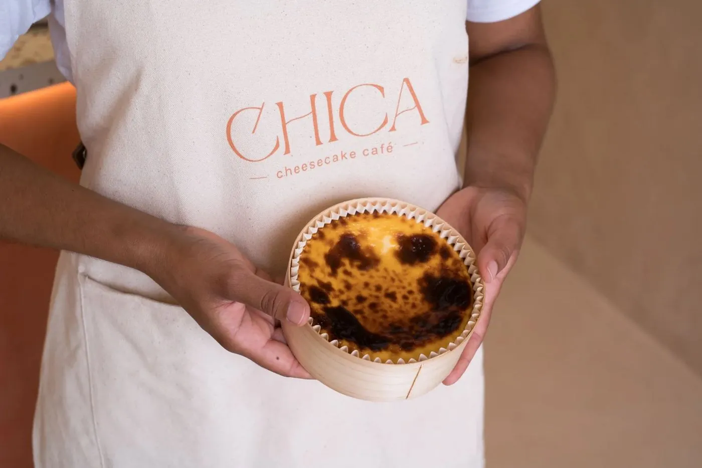 Una cheesecake de CHICA recién horneada en su caja de madera, sostenida por alguien con el delantal de CHICA